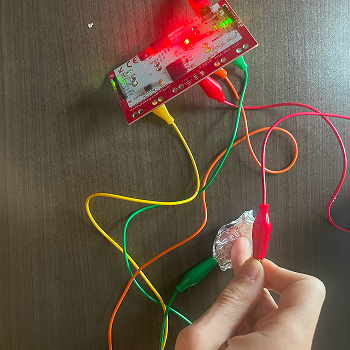 Testing aluminum foil as a conductive surface, cutting a small piece, connecting a clip, and touching it to compare its conductivity and feel to copper tape and graphite.